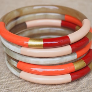 Könnte beinhalten: Ein Stapel von vier bunten Armreifen mit einem roten, goldenen und pfirsichfarbenen Design. Die Armreifen sind aus einem natürlichen Material gefertigt und haben eine glänzende Oberfläche.