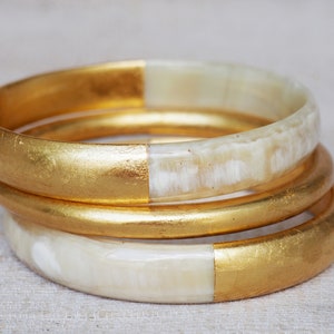 Puede incluir: Tres brazaletes dorados y blancos apilados uno encima del otro. Los brazaletes están hechos de un material blanco con un acabado dorado en el exterior.