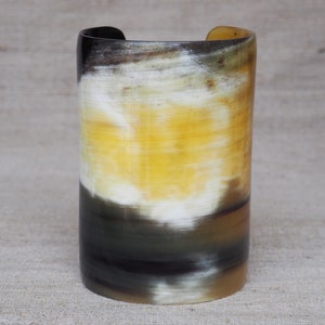 May include: A cylindrical cuff bracelet made from natural horn. The bracelet features a gradient of colors, including black, white, yellow, and brown. The cuff is approximately 2.5 inches in height and has a smooth, polished finish. The bracelet is a unique accessory.