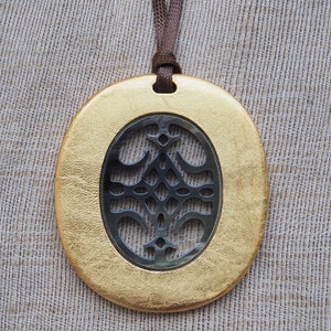 May include: A gold oval pendant with a cutout design in the center. The cutout design is a black and silver geometric pattern. The pendant is hanging from a brown cord.