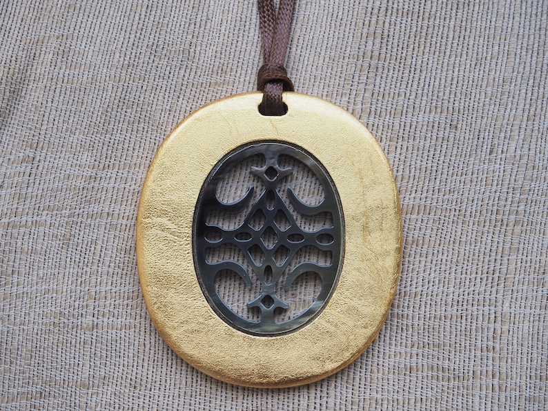 May include: A gold oval pendant with a cutout design in the center. The cutout design is a black and silver geometric pattern. The pendant is hanging from a brown cord.