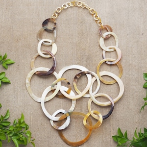 Puede incluir: Un collar hecho de múltiples anillos conectados de cuerno marrón y blanco. Los anillos son de diferentes tamaños y están conectados por una cadena dorada.