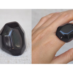 Könnte beinhalten: Ein großer, schwarzer, geometrischer Ring aus Harz. Der Ring ist in Form eines facettierten Edelsteins geformt und hat eine hochglänzende Oberfläche.