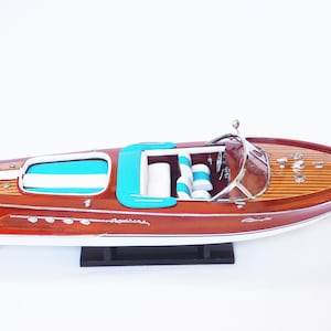 Op de afbeelding: Een houten model van een klassieke mahonie speedboot met een witte romp en turquoise interieur. De boot staat op een zwarte standaard en heeft de naam "Aquarama" aan de zijkant.