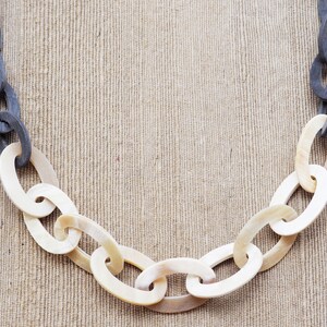 May include: A chunky chain necklace with alternating links of dark gray and off-white oval shapes. The necklace is displayed against a neutral, textured background, highlighting the contrast in colors and textures.