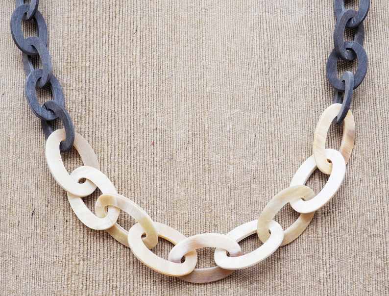 May include: A chunky chain necklace with alternating links of dark gray and off-white oval shapes. The necklace is displayed against a neutral, textured background, highlighting the contrast in colors and textures.