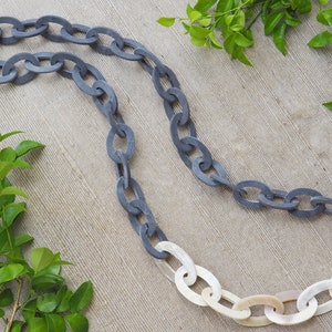 May include: A gray and white chain necklace with oval links. The necklace is made of a dark gray material that transitions to white links. The necklace is displayed on a neutral background with green foliage.