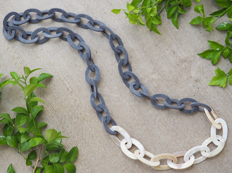 May include: A gray and white chain necklace with oval links. The necklace is made of a dark gray material that transitions to white links. The necklace is displayed on a neutral background with green foliage.