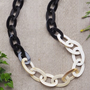 May include: A chunky chain necklace featuring large oval links. The necklace alternates between black and off-white links, creating a bold contrast. The jewelry is displayed on a neutral background, highlighting the necklace's design.