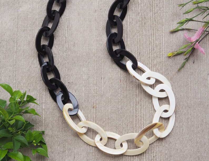 May include: A chunky chain necklace featuring large oval links. The necklace alternates between black and off-white links, creating a bold contrast. The jewelry is displayed on a neutral background, highlighting the necklace's design.