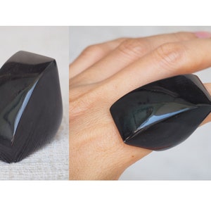 Könnte beinhalten: Ein großer, schwarzer, geometrischer Ring mit einer glatten, polierten Oberfläche. Der Ring ist in Form eines abgerundeten Dreiecks und hat eine leicht gewölbte Oberseite.