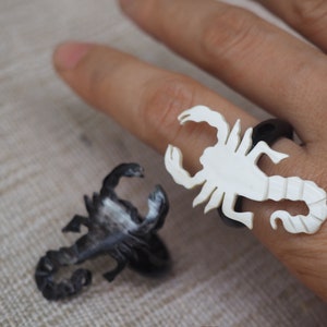Könnte beinhalten: Zwei Skorpionringe, einer schwarz und einer weiß. Der weiße Ring wird an einem Finger getragen.