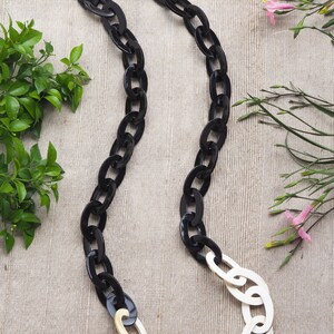 May include: A long, oval-link necklace with a striking contrast. The upper portion features glossy black links, transitioning to cream-colored links at the bottom. The necklace is displayed on a neutral linen surface, with greenery and pink flowers in the background.