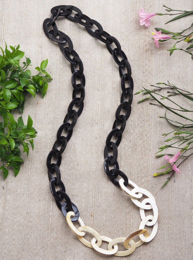 May include: A long, oval-link necklace with a striking contrast. The upper portion features glossy black links, transitioning to cream-colored links at the bottom. The necklace is displayed on a neutral linen surface, with greenery and pink flowers in the background.