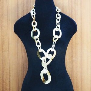 以下が含まれることがあります： 大きな楕円形のリンクで作られた、長く、白く、ずんぐりしたチェーンネックレス。ネックレスは黒いマネキンに飾られています。