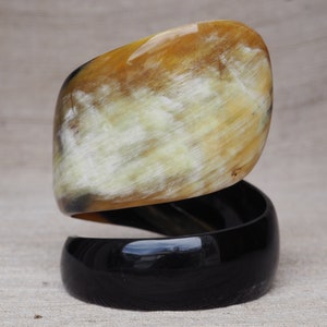 Op de afbeelding: Een paar armbanden, een is een brede, gebogen armband van geel en wit hoorn, de andere is een zwarte hoorn armband.