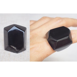 Könnte beinhalten: Ein schwarzer, facettierter geometrischer Ring. Der Ring ist groß und hat ein einzigartiges, modernes Design.
