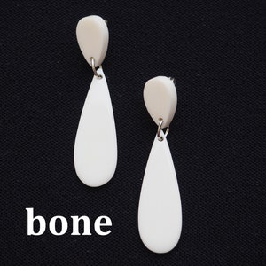 以下が含まれることがあります： 小さな丸みを帯びた上部が付いた、象牙色のしずく型のイヤリングのペア。イヤリングは銀色の金属ループで接続されています。画像の下部には「bone」という単語が白で印刷されています。