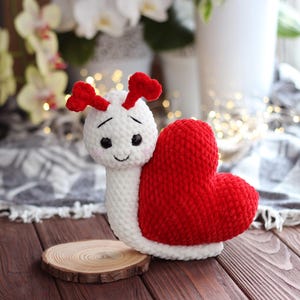 Può includere: Una lumaca bianca all'uncinetto con un guscio a forma di cuore rosso. La lumaca ha un viso sorridente con occhi neri e un naso rosso.