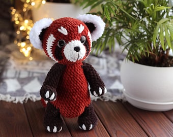 May include: A handmade crochet red panda plush toy. It features a red body, dark brown limbs, and white accents on the ears and face. The toy is positioned on a wooden surface.