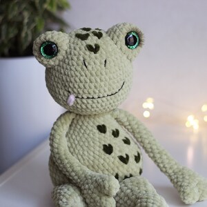 May include: Green crocheted frog plush toy with large green eyes and a pink tongue sticking out. The frog has a smiling expression and is sitting on a white surface.