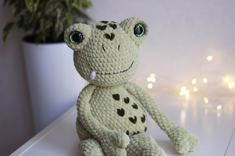 May include: Green crocheted frog plush toy with large green eyes and a pink tongue sticking out. The frog has a smiling expression and is sitting on a white surface.