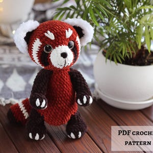 Könnte beinhalten: Handgehäkeltes rotes Panda-Plüschtier mit weißen Akzenten an Ohren, Gesicht und Pfoten. Das Spielzeug hat einen dunkelbraunen Körper und Schwanz und schwarze Augen. Der Text "PDF CROCHET PATTERN" ist sichtbar.
