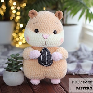 Könnte beinhalten: Ein gehäkeltes Plüsch-Hamster-Spielzeug mit hellbraunem Fell, weißer Schnauze, rosa Wangen und kleinen Ohren. Der Hamster hat schwarze Augen, eine rosa Nase und hellrosa Pfoten. Er hält einen dunkelgrauen Sonnenblumenkern mit weißen Nähten.