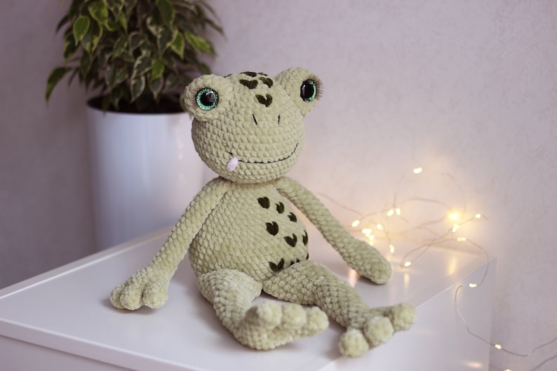 May include: A green crocheted frog plush toy with black spots and large green eyes. The frog is sitting on a white surface.