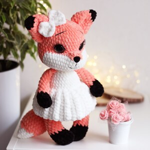May include: A crocheted plush fox toy with a white dress and a white bow. The fox has black eyes and a black nose. The toy is standing on a white surface.