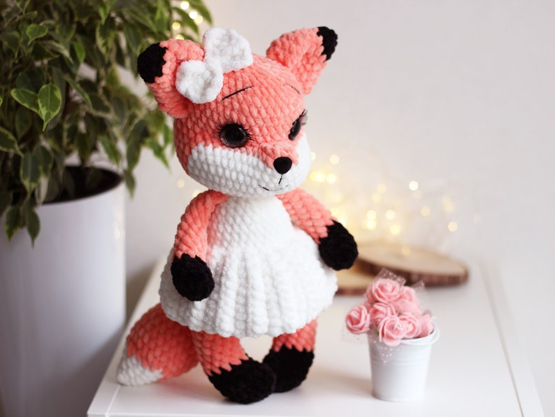 May include: A crocheted plush fox toy with a white dress and a white bow. The fox has black eyes and a black nose. The toy is standing on a white surface.