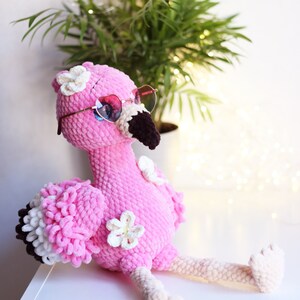 May include: Pink crocheted flamingo plush toy wearing heart-shaped sunglasses and white flower embellishments. The flamingo is sitting on a white surface.