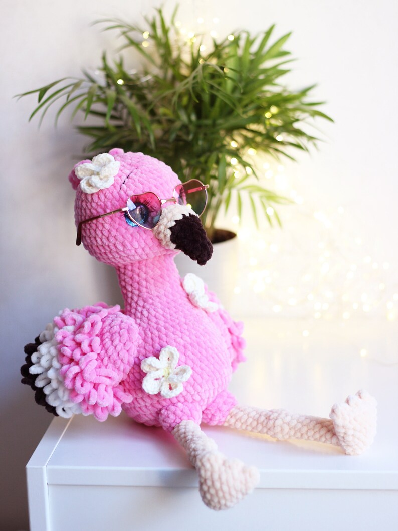 May include: Pink crocheted flamingo plush toy wearing heart-shaped sunglasses and white flower embellishments. The flamingo is sitting on a white surface.