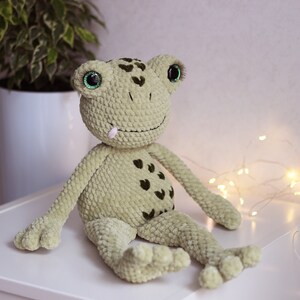 May include: A green crocheted frog plush toy with black spots and large green eyes. The frog is sitting on a white surface.