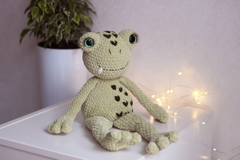 May include: A green crocheted frog plush toy with black spots and large green eyes. The frog is sitting on a white surface.