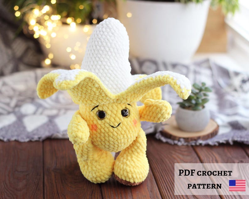 May include: A handmade crochet banana plushie with a smiling face, rosy cheeks, and brown feet. The banana peel is yellow with white accents, and the banana itself is white. The image includes the text "PDF CROCHET PATTERN" and the American flag.