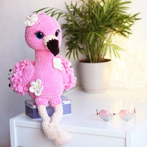 May include: Pink crocheted flamingo toy with white flowers and a brown beak. The flamingo is sitting on a white surface with a small tin box and heart-shaped sunglasses.