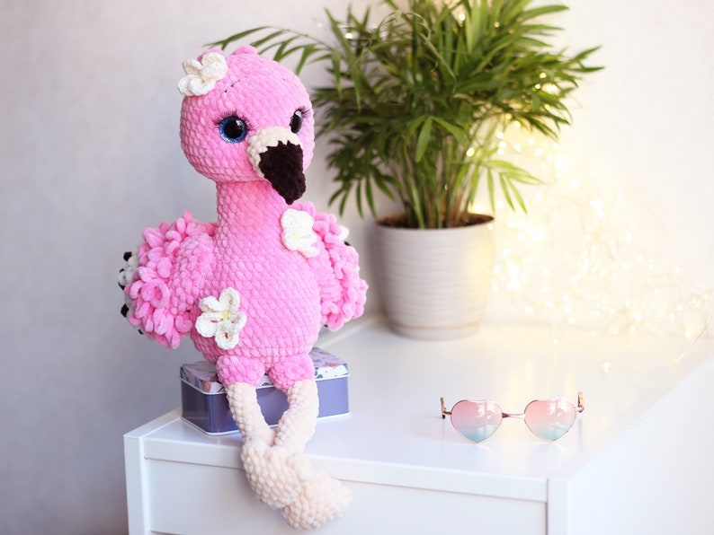 May include: Pink crocheted flamingo toy with white flowers and a brown beak. The flamingo is sitting on a white surface with a small tin box and heart-shaped sunglasses.