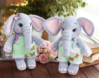 Può includere: Due peluche a forma di elefante fatti a mano all'uncinetto. Gli elefanti sono azzurri con orecchie foderate di rosa e indossano salopette verdi e a righe pastello. Ognuno ha un dettaglio a farfalla e un fiocco colorato.