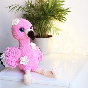 May include: Pink crocheted flamingo plush toy with white flowers and black eyes. The flamingo is sitting on a white surface.