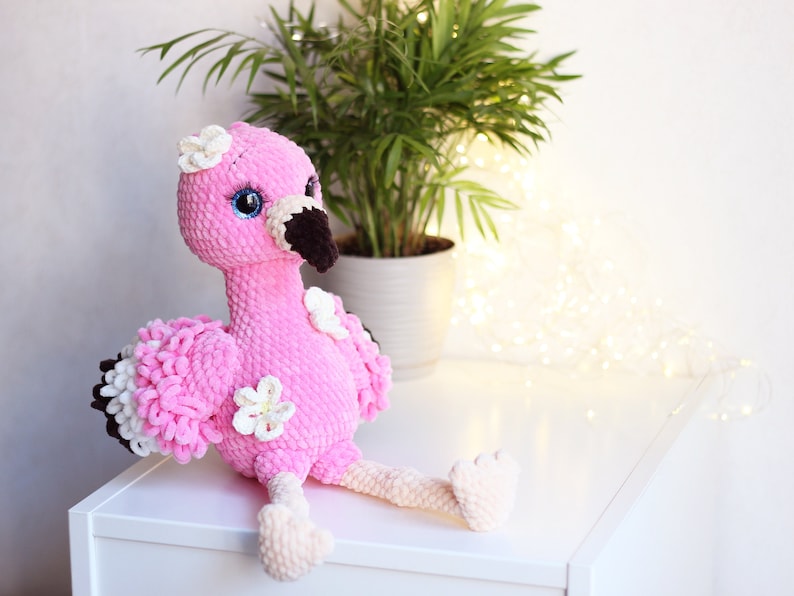May include: Pink crocheted flamingo plush toy with white flowers and black eyes. The flamingo is sitting on a white surface.