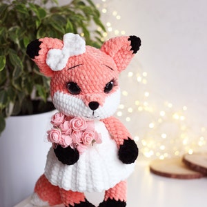 May include: A crocheted fox plush toy wearing a white dress and holding a bouquet of pink roses. The fox has black eyes, a white bow, and black accents on its ears, paws, and tail.