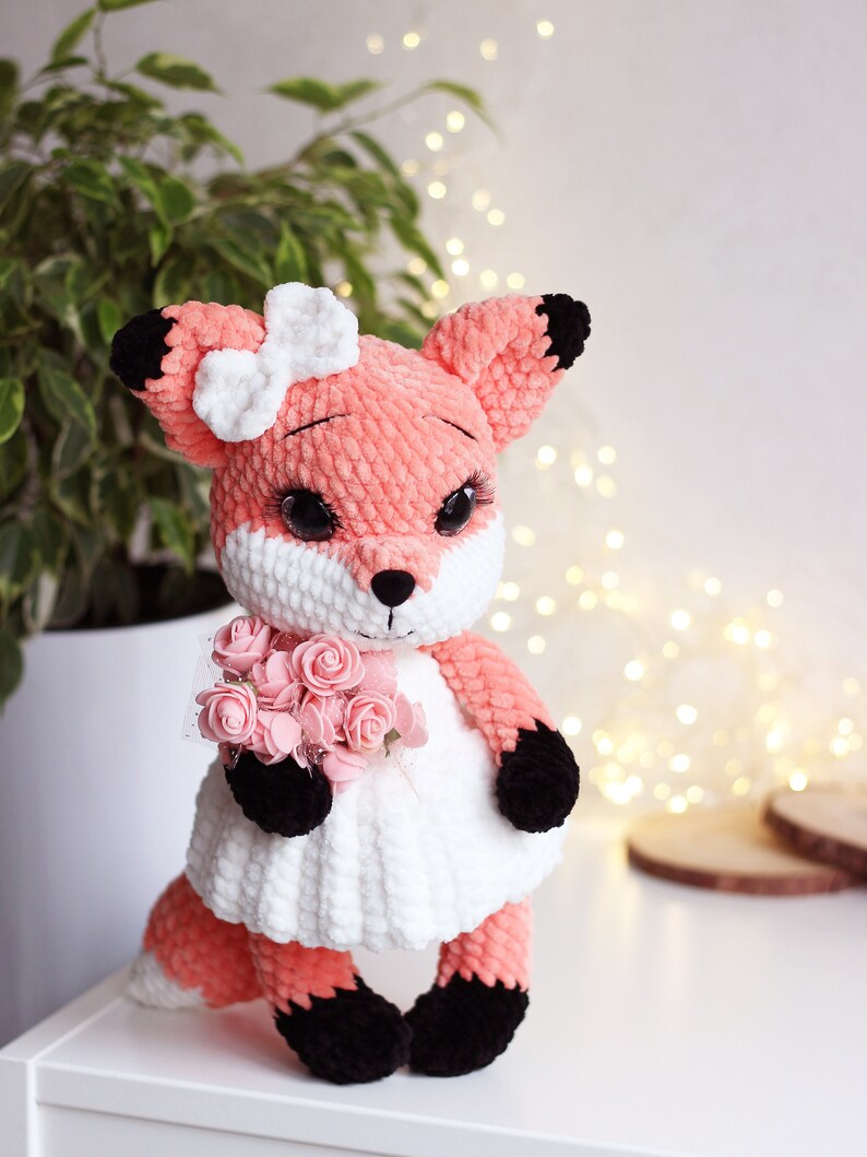 May include: A crocheted fox plush toy wearing a white dress and holding a bouquet of pink roses. The fox has black eyes, a white bow, and black accents on its ears, paws, and tail.