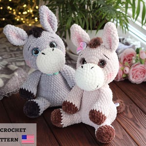 May include: Two handmade crochet donkey plushies. One is gray with black and brown accents, the other is pink with brown accents. Both have large ears, white muzzles, and embroidered eyes. The plushies are sitting on a wooden surface.