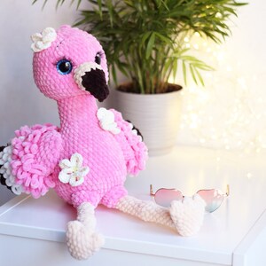 May include: A pink crocheted flamingo toy with white flowers and blue eyes. The flamingo is sitting on a white surface with a pair of pink sunglasses.