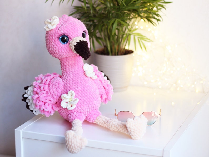 May include: A pink crocheted flamingo toy with white flowers and blue eyes. The flamingo is sitting on a white surface with a pair of pink sunglasses.