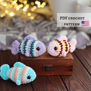 Op de afbeelding: Drie kleine, gehaakte pluche vissen met zwarte veiligheidsoogjes en roze wangen. Eén vis is paars, groenblauw en wit; één is geel, rood en wit; één is blauw en perzikkleurig. Tekst: PDF CROCHET PATTERN met een Amerikaanse vlag.