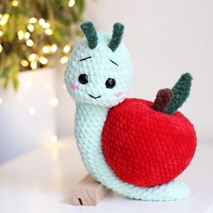 Può includere: Una lumaca di uncinetto verde con una mela di uncinetto rossa sulla schiena. La lumaca ha un viso sorridente con occhi neri e guance rosa.