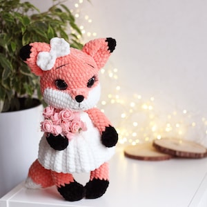May include: A crocheted fox plush toy wearing a white dress and holding a bouquet of pink flowers. The fox has black eyes, a pink nose, and black ears.