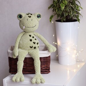May include: A green crocheted frog with a pink tongue sitting in a brown wicker basket. The frog has green eyes and a pattern of dark green hearts on its belly.